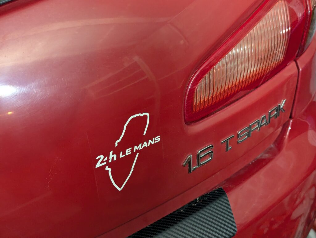 Close-up of the trunk lid of a red Alfa Romeo 147 1.6 TS sporting a white decal showing the outline of the Circuit de la Sarthe with the text “24h Le Mans”.