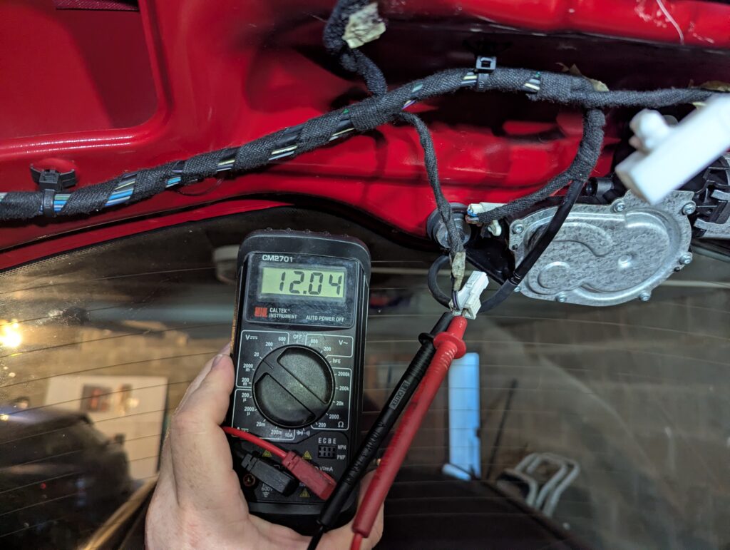 Inside view of the raised trunk lid of my Alfa Romeo 147 with the lining removed. The trunk lock is disconnected and black and red multimeter probes are inserted into the plug. I am holding the multimeter in my left hand; it is set to 20 V and the display is showing12.04.