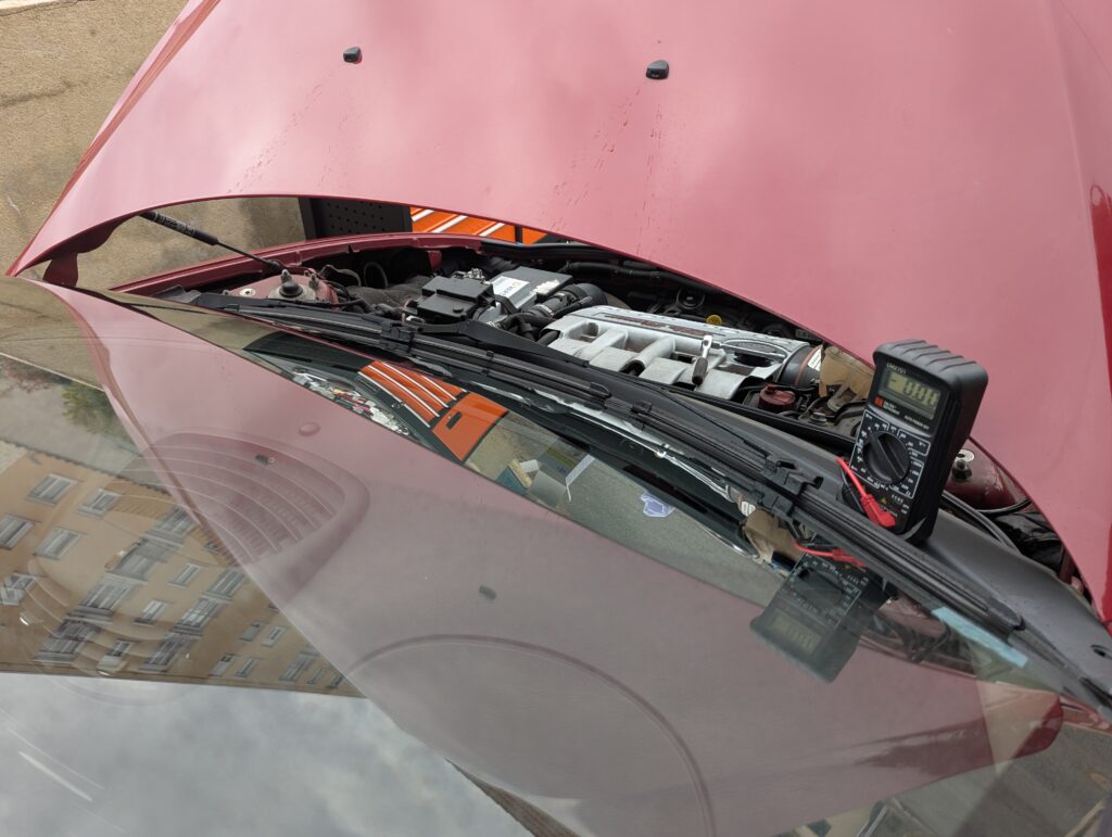 View of the front of a red Alfa Romeo 147 from right above / next to the passenger window. The hood is open and there are traces of washer fluid on it. A multimeter is propped up against the hood so that it is visible from the driver's seat. A ratchet is lying on top of the engine, which is visible through the gap between the rear edge of the open hood and the body.